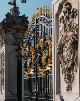 Ornate iron gates with intricate golden embellishments. The design includes detailed sculptures and decorative patterns. Stone structures with elaborate carvings flank the gates, depicting various symbols and figures.