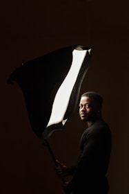 A person is standing in a dimly lit space, holding a large studio light with a softbox. The light illuminates their face and part of their body, creating a dramatic contrast against the dark background.