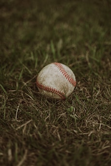 A worn-out baseball lies on a patch of vibrant green grass, surrounded by small blades and patches of earth.