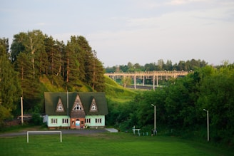 A quaint green house with a triangular roof and multiple windows sits in a lush, verdant landscape. Tall, dense trees rise behind the house, while a football goalpost stands on the green field in front. In the background, a bridge stretches across green hills, surrounded by a vast expanse of trees under a bright, clear sky.