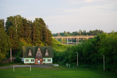 A quaint green house with a triangular roof and multiple windows sits in a lush, verdant landscape. Tall, dense trees rise behind the house, while a football goalpost stands on the green field in front. In the background, a bridge stretches across green hills, surrounded by a vast expanse of trees under a bright, clear sky.