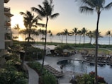 A coastal resort scene featuring a luxurious swimming pool surrounded by lounge chairs and palm trees. Guests relax by the poolside under shaded umbrellas. The ocean is visible in the background, reflecting the golden hues of a setting sun. Buildings and lush greenery frame the scene, enhancing the tropical atmosphere.