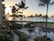 A coastal resort scene featuring a luxurious swimming pool surrounded by lounge chairs and palm trees. Guests relax by the poolside under shaded umbrellas. The ocean is visible in the background, reflecting the golden hues of a setting sun. Buildings and lush greenery frame the scene, enhancing the tropical atmosphere.