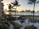 A coastal resort scene featuring a luxurious swimming pool surrounded by lounge chairs and palm trees. Guests relax by the poolside under shaded umbrellas. The ocean is visible in the background, reflecting the golden hues of a setting sun. Buildings and lush greenery frame the scene, enhancing the tropical atmosphere.