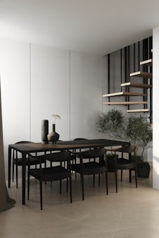 A modern, minimalist dining area featuring a sleek dark wood table with matching chairs. Two vases with a simple design sit on the table. The background includes a white wall and a floating wooden staircase with black railings. Potted plants are placed on the floor, adding a touch of greenery.