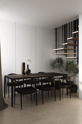 A modern, minimalist dining area featuring a sleek dark wood table with matching chairs. Two vases with a simple design sit on the table. The background includes a white wall and a floating wooden staircase with black railings. Potted plants are placed on the floor, adding a touch of greenery.