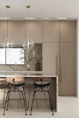 A modern kitchen features a sleek island with a polished marble countertop. Two bar stools with wooden seats and black metal legs are placed at the island. Hanging above the island are two minimalist pendant lights with clear spherical shades. The background includes a large window, beige cabinetry with vertical handles, and a decorative cutting board with a vase containing green foliage.