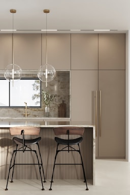 A modern kitchen features a sleek island with a polished marble countertop. Two bar stools with wooden seats and black metal legs are placed at the island. Hanging above the island are two minimalist pendant lights with clear spherical shades. The background includes a large window, beige cabinetry with vertical handles, and a decorative cutting board with a vase containing green foliage.