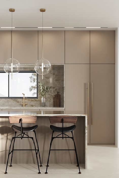 A modern kitchen features a sleek island with a polished marble countertop. Two bar stools with wooden seats and black metal legs are placed at the island. Hanging above the island are two minimalist pendant lights with clear spherical shades. The background includes a large window, beige cabinetry with vertical handles, and a decorative cutting board with a vase containing green foliage.