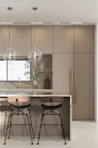 A modern kitchen features a sleek island with a polished marble countertop. Two bar stools with wooden seats and black metal legs are placed at the island. Hanging above the island are two minimalist pendant lights with clear spherical shades. The background includes a large window, beige cabinetry with vertical handles, and a decorative cutting board with a vase containing green foliage.
