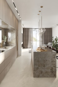 A modern kitchen interior with sleek cabinetry and a long marble-topped island. Pendant lights hang above the island, which features a tray with bread and glasses. Large windows with sheer curtains allow natural light to fill the space, accentuating the minimalist design and neutral color palette. Several potted plants add a touch of greenery.