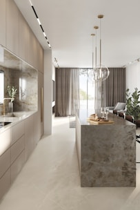 A modern kitchen interior with sleek cabinetry and a long marble-topped island. Pendant lights hang above the island, which features a tray with bread and glasses. Large windows with sheer curtains allow natural light to fill the space, accentuating the minimalist design and neutral color palette. Several potted plants add a touch of greenery.