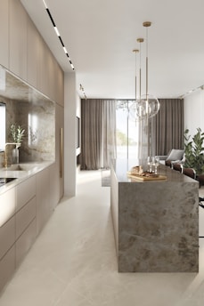 A modern kitchen interior with sleek cabinetry and a long marble-topped island. Pendant lights hang above the island, which features a tray with bread and glasses. Large windows with sheer curtains allow natural light to fill the space, accentuating the minimalist design and neutral color palette. Several potted plants add a touch of greenery.