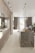 A modern kitchen interior with sleek cabinetry and a long marble-topped island. Pendant lights hang above the island, which features a tray with bread and glasses. Large windows with sheer curtains allow natural light to fill the space, accentuating the minimalist design and neutral color palette. Several potted plants add a touch of greenery.