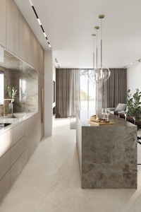 A modern kitchen interior with sleek cabinetry and a long marble-topped island. Pendant lights hang above the island, which features a tray with bread and glasses. Large windows with sheer curtains allow natural light to fill the space, accentuating the minimalist design and neutral color palette. Several potted plants add a touch of greenery.