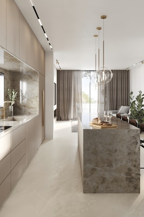 A modern kitchen interior with sleek cabinetry and a long marble-topped island. Pendant lights hang above the island, which features a tray with bread and glasses. Large windows with sheer curtains allow natural light to fill the space, accentuating the minimalist design and neutral color palette. Several potted plants add a touch of greenery.