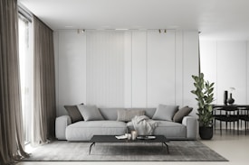 A modern living room featuring a large gray sectional sofa adorned with various pillows in shades of gray and beige. In front of the sofa is a sleek, low-profile black coffee table with decorative items including candles and a small book. A textured gray rug is laid out on the floor beneath the table. Tall, elegant curtains in a muted tone frame large windows on the left side, allowing natural light to softly illuminate the room. A potted plant adds a touch of greenery next to the sofa, blending well with the minimalist decor. In the background, a small dining area can be seen with a round table and black chairs, along with a few vases on a side table.