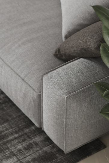 A detailed view of a gray upholstered sofa with a textured surface, accompanied by two cushions in different shades of gray. A plant with broad green leaves is partially visible on the right side, and the sofa is placed atop a dark, patterned rug.