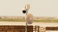 A security camera and satellite dish mounted on a pole, positioned on a brick wall. The scene overlooks a flat, rural landscape with sparse vegetation in the distance, under a clear sky.