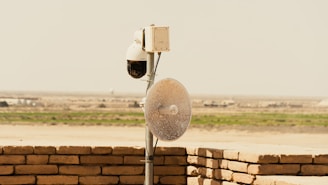 A security camera and satellite dish mounted on a pole, positioned on a brick wall. The scene overlooks a flat, rural landscape with sparse vegetation in the distance, under a clear sky.