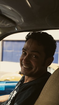 A man sitting in the driver's seat of a vehicle, smiling warmly. The interior of the car is visible, with a beige seat cover and a glimpse of the car's blue exterior visible through the window.