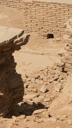 Ancient ruins featuring exposed brickwork and scattered stones surround a partially intact wall. The scene is set in a dusty environment with signs of erosion and decay, indicating a historical or archaeological site.