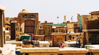 A cemetery with numerous tombstones and grave markers featuring Arabic script. The structures are made of stone and brick, exhibiting various shades of brown and beige. Some grave markers have decorations including flowers and small bowls. The scene appears to be under clear daylight.