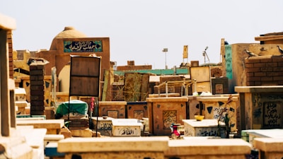 A cemetery with numerous tombstones and grave markers featuring Arabic script. The structures are made of stone and brick, exhibiting various shades of brown and beige. Some grave markers have decorations including flowers and small bowls. The scene appears to be under clear daylight.