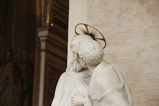 A detailed marble sculpture depicts a religious figure with curly hair and a halo, suggesting holiness or sainthood. The background features intricate architectural design and a visible section of a mural or painting, hinting at a sacred or significant historical context.