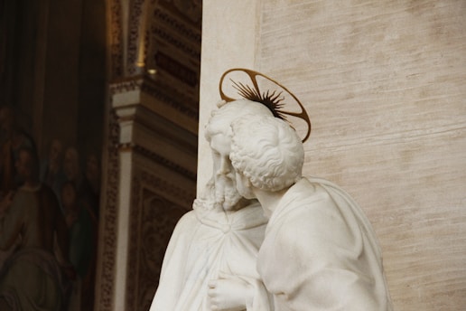 A detailed marble sculpture depicts a religious figure with curly hair and a halo, suggesting holiness or sainthood. The background features intricate architectural design and a visible section of a mural or painting, hinting at a sacred or significant historical context.