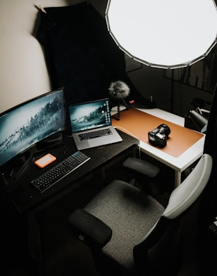 A workspace featuring a computer setup with a keyboard, a laptop, and a large curved monitor displaying a serene landscape. A camera sits on a side table beside a bright softbox light, providing illumination. The space includes a chair positioned at the desk and an external hard drive near the keyboard.