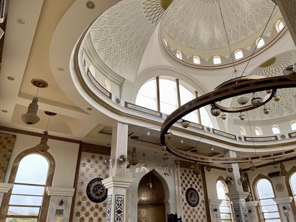 The image depicts an intricately designed interior of a building, featuring a large domed ceiling with patterned detailing and circular windows. There are grand chandeliers hanging from the ceiling, and decorative elements such as ornate pillars and Arabic calligraphy adorn the walls. The space is well-lit with natural light coming through large arched windows.