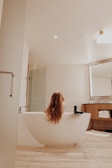 A modern bathroom features a sleek, freestanding bathtub with a person relaxing inside, their long hair cascading over the tub's edge. The room is elegantly designed with neutral tones, wooden cabinet drawers filled with neatly stacked towels, and a large wall mirror. A glass shower stall is positioned next to the bathtub, and soft lighting adds a warm ambiance.