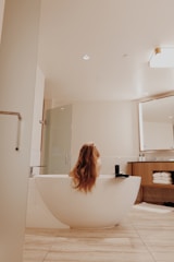 A modern bathroom features a sleek, freestanding bathtub with a person relaxing inside, their long hair cascading over the tub's edge. The room is elegantly designed with neutral tones, wooden cabinet drawers filled with neatly stacked towels, and a large wall mirror. A glass shower stall is positioned next to the bathtub, and soft lighting adds a warm ambiance.
