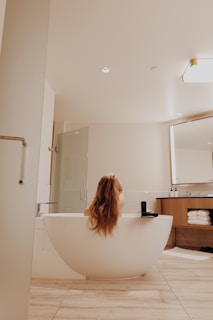A modern bathroom features a sleek, freestanding bathtub with a person relaxing inside, their long hair cascading over the tub's edge. The room is elegantly designed with neutral tones, wooden cabinet drawers filled with neatly stacked towels, and a large wall mirror. A glass shower stall is positioned next to the bathtub, and soft lighting adds a warm ambiance.