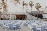 An outdoor event setup features numerous round tables covered with blue and white patterned tablecloths, each surrounded by white folding chairs. String lights are hung above, creating a warm, festive ambiance. Tall palm trees and a stone wall provide a picturesque backdrop, and people can be seen preparing the space.