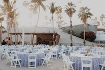 An outdoor event setup features numerous round tables covered with blue and white patterned tablecloths, each surrounded by white folding chairs. String lights are hung above, creating a warm, festive ambiance. Tall palm trees and a stone wall provide a picturesque backdrop, and people can be seen preparing the space.