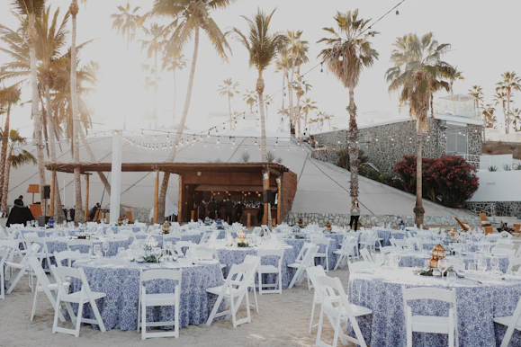 An outdoor event setup features numerous round tables covered with blue and white patterned tablecloths, each surrounded by white folding chairs. String lights are hung above, creating a warm, festive ambiance. Tall palm trees and a stone wall provide a picturesque backdrop, and people can be seen preparing the space.