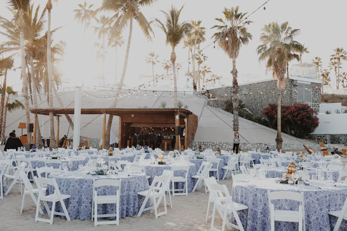 An outdoor event setup features numerous round tables covered with blue and white patterned tablecloths, each surrounded by white folding chairs. String lights are hung above, creating a warm, festive ambiance. Tall palm trees and a stone wall provide a picturesque backdrop, and people can be seen preparing the space.