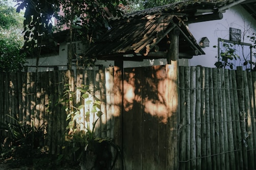A rustic wooden fence with bamboo poles and a traditional wooden gate partially shaded by overhanging tree branches. Sunlight filters through the leaves creating dappled patterns on the fence. Green foliage surrounds the area adding an overgrown and natural atmosphere.