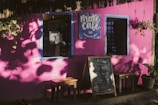 A vibrant pink wall serves as the backdrop for a cozy café, adorned with leafy shadows. The café's name, Maré Café, is prominently displayed on a blue circular sign with white lettering. Two small windows with blue frames enhance the wall's color, and a menu board rests on the ground near a wooden stool. Potted plants and hanging baskets add a touch of greenery to the scene, reflecting a warm and inviting atmosphere.
