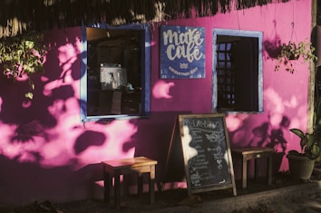 A vibrant pink wall serves as the backdrop for a cozy café, adorned with leafy shadows. The café's name, Maré Café, is prominently displayed on a blue circular sign with white lettering. Two small windows with blue frames enhance the wall's color, and a menu board rests on the ground near a wooden stool. Potted plants and hanging baskets add a touch of greenery to the scene, reflecting a warm and inviting atmosphere.