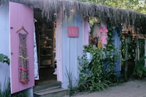 A quaint beachside boutique with lavender-painted wooden walls, adorned with a floral dress and a pink bikini displayed outside. The shop entrance is flanked by lush greenery and a straw-thatched roof, creating a tropical ambiance. Inside, various garments and accessories are neatly arranged on shelves.