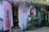 A quaint beachside boutique with lavender-painted wooden walls, adorned with a floral dress and a pink bikini displayed outside. The shop entrance is flanked by lush greenery and a straw-thatched roof, creating a tropical ambiance. Inside, various garments and accessories are neatly arranged on shelves.