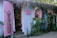 A quaint beachside boutique with lavender-painted wooden walls, adorned with a floral dress and a pink bikini displayed outside. The shop entrance is flanked by lush greenery and a straw-thatched roof, creating a tropical ambiance. Inside, various garments and accessories are neatly arranged on shelves.