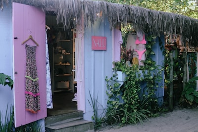 A quaint beachside boutique with lavender-painted wooden walls, adorned with a floral dress and a pink bikini displayed outside. The shop entrance is flanked by lush greenery and a straw-thatched roof, creating a tropical ambiance. Inside, various garments and accessories are neatly arranged on shelves.