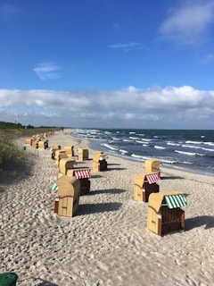 A sandy beach with rows of wicker beach chairs featuring striped canopies, positioned along the shoreline. The beach is bordered by the sea with gentle waves, under a blue sky with scattered clouds. Grassy dunes and a wind turbine are visible in the background.