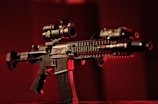 A black rifle with various attachments, including a scope and other tactical gear, is set against a dark, red-tinged background. The lighting emphasizes the structure of the weapon, casting shadows and highlighting its details.