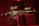 A black rifle with various attachments, including a scope and other tactical gear, is set against a dark, red-tinged background. The lighting emphasizes the structure of the weapon, casting shadows and highlighting its details.