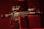A black rifle with various attachments, including a scope and other tactical gear, is set against a dark, red-tinged background. The lighting emphasizes the structure of the weapon, casting shadows and highlighting its details.